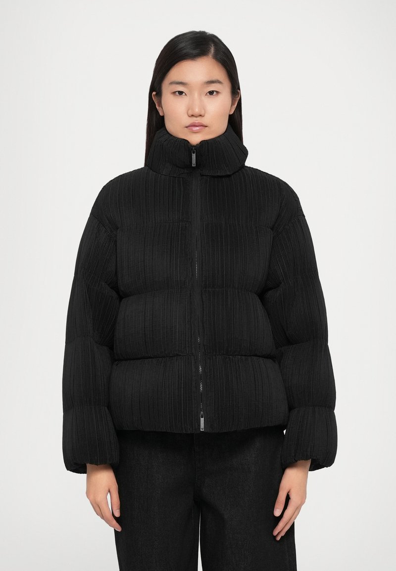 Black puffer jacket with a high collar, ribbed texture, front zipper, and boxy silhouette. Paired with dark jeans.