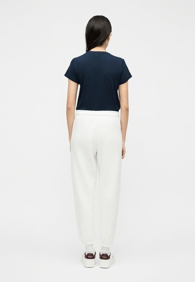 Γυναίκα που στέκεται με την πλάτη, φορώντας μπλε μπλουζάκι, λευκή φόρμα, λευκές κάλτσες και λευκά αθλητικά παπούτσια με σκούρα τακούνια.