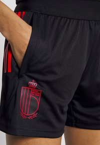 Czarne spodenki sportowe z czerwonymi pionowymi paskami, z logo przedstawiającym koronę i napisem "ROYAL BELGIAN FA 1895" po lewej stronie.