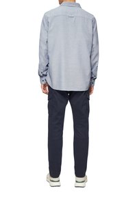 Homme se tenant de dos, portant une chemise à boutons bleu clair, un pantalon cargo noir et des baskets blanches avec des accents verts.