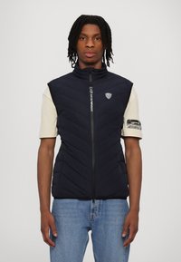 Navy quiltet ermeløs vest med høy krage, glidelås foran og logo detaljer. Båret over en lysfarget t-skjorte med korte ermer og jeans.