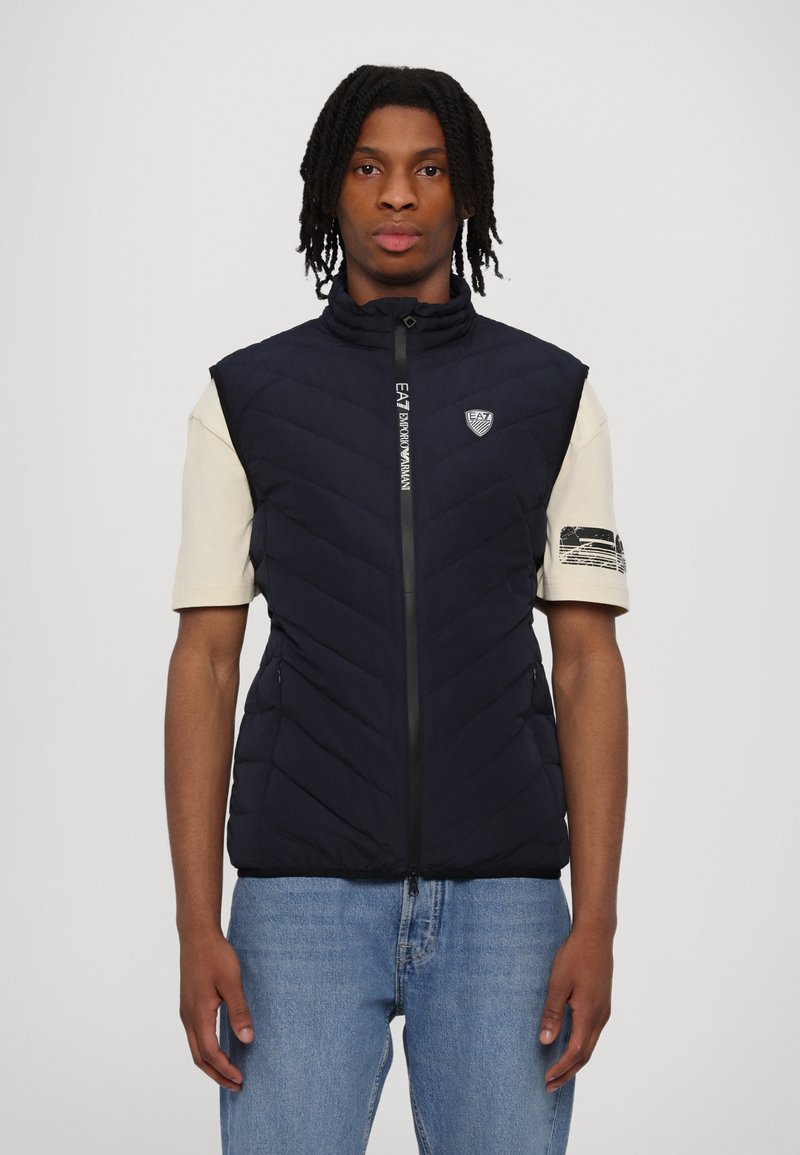 Navy quiltet ermeløs vest med høy krage, glidelås foran og logo detaljer. Båret over en lysfarget t-skjorte med korte ermer og jeans.