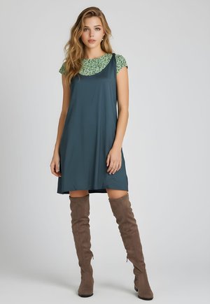 Mujer con cabello largo y rubio, lleva una camisa verde con estampado debajo de un vestido de tirantes verde azulado oscuro y botas marrones hasta la rodilla, de pie contra un fondo gris.