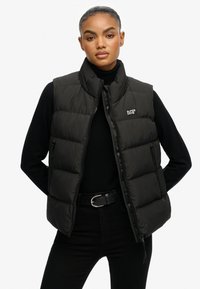 Chaleco acolchado negro con cremallera frontal, material suave, cuello alto y logo en parche. Llevado sobre un suéter negro y combinado con pantalones negros.