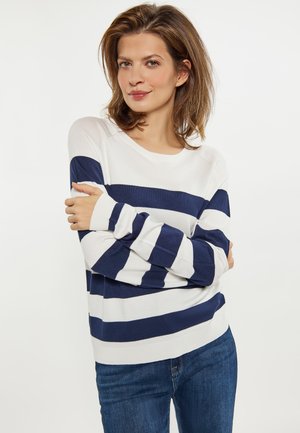 Kvinde med lyst brunt hår iført en hvid sweater med brede marineblå vandrette striber og blå jeans, der står med armene krydset.