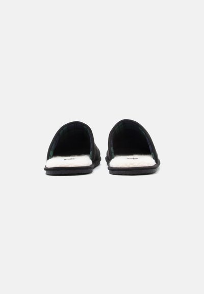 Melnu slēpjamo čību pāris ar pārklājuma audumu un baltām pūkainām zolēm, redzams no priekšpuses uz gaiša fona.