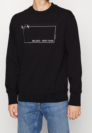 Homme portant un sweat-shirt noir Armani Exchange avec le texte « MILANO - NEW YORK » et un jean bleu, debout devant un fond uni.