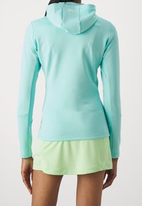 Dlouhá mikina s kapucí v světlemodré látce, s hladkým povrchem. Spárováno s krátkou sukní v mátově zelené barvě, která má vrstvený design.