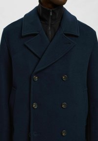 Cappotto doppiopetto in lana blu navy con ampi risvolti, sei bottoni rotondi e tasche laterali. Presenta uno strato interno nero con zip.