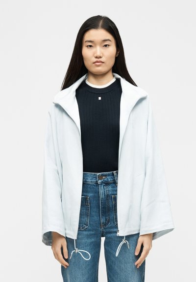 Γυναίκα με μακριά σκούρα μαλλιά, φορώντας ανοιχτό μπλε μπουφάν με φερμουάρ πάνω από μαύρο ριμπ μπλουζάκι και ψηλόμεσα μπλε τζιν, σε λευκό φόντο.