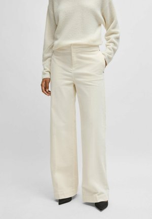 Weite Hose aus cremefarbenem Stoff, mit Seitentaschen und einer glatten Textur. Kombiniert mit einem leichten Strickpullover und schwarzen spitzen Absätzen.