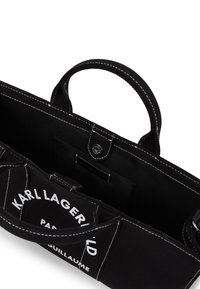 Bolsa de tote negra de lona con costuras blancas y logo. Asas superiores, interior abierto, bolsillo y diseño estructurado.