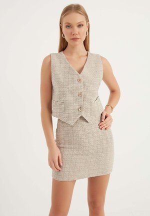 Beige Tweed-Weste mit V-Ausschnitt, ausgestattet mit drei vorderen Knöpfen und seitlichen Taschen. Kombiniert mit einem figurbetonten Tweed-Minirock.