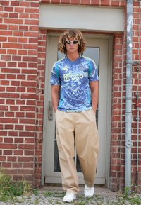 Maglietta sportiva a fantasia blu e verde con logo "Ziggo", pantaloni cargo beige e scarpe da ginnastica bianche, accessoriate con occhiali da sole bianchi.