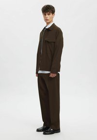 Ruda užsegama švarkelio tipo striukė su plokščiu apykakliu ir krūtinės kišene, derančiomis rudos spalvos kelnėmis ir juodais blizgiais batais.