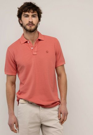 Hombre con cabello rizado que lleva una camisa tipo polo color salmón y pantalones beige claro, de pie frente a un fondo liso.