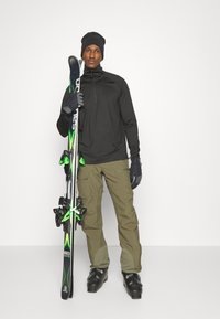 Top de manga larga negro, pantalones de esquí verde oliva, guantes negros y botas de esquí. Sosteniendo esquís negros y verdes con fijaciones, de pie sobre un fondo blanco.