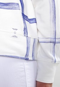 Suéter blanco con patrón de rayas azules, textura suave y etiqueta de marca. Detalle de costura visible a lo largo del dobladillo y las mangas.
