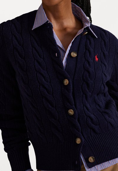 Woman wearing navy blue cable-knit cardigan with brown buttons over blue and red striped collared shirt.