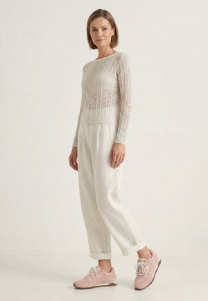 Woman standing wearing a beige knit sweater, loose white pants cuffed at ankles, and pink lace-up sneakers on a neutral background.