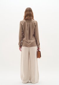 Brown satin blouse with buttoned cuffs, back pleat, paired with loose white linen trousers and a small brown handbag. Minimalist design.