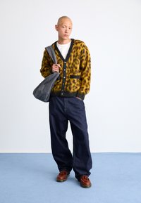 Κίτρινο και μαύρο καρό πουλόβερ με κουμπιά, γκρι τσάντα ώμου, λευκό πουκάμισο, σκούρα μπλε φαρδιά τζιν παντελόνια, καφέ παπούτσια.