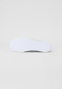 Bílá podrážka tenisky s texturovaným povrchem, obsahující horizontální drážky a branding. Zaoblená špička a plochý design vhodný pro volný čas.