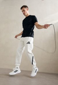 Młody mężczyzna w czarnej koszulce i białych spodniach Adidas skacze na skakance wewnątrz pomieszczenia na tle gładkiej beżowej ściany.