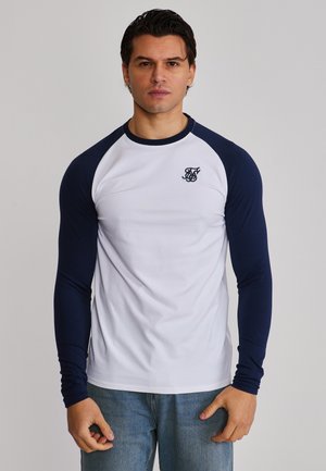 Młody mężczyzna ubrany w białą koszulę z długim rękawem, granatowymi rękawami i małym logo na piersi, stojący na jasnym, jednolitym tle.