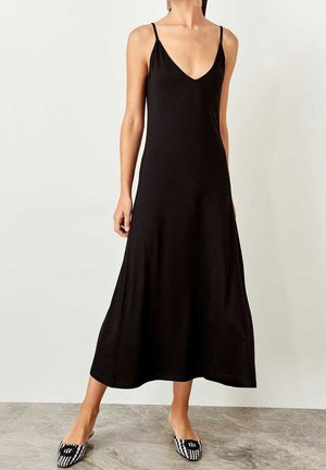 Frau trägt ein ärmelloses schwarzes Midi-Kleid mit dünnen Trägern und V-Ausschnitt, kombiniert mit schwarz-weißen karierter spitz zulaufenden Ballerinas.