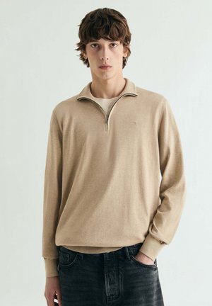 Jersey beige de manga larga con cuello de media cremallera, hecho de material suave, con puños acanalados, combinado con vaqueros oscuros.