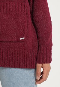 Burgundská oversized pletená svetr s žebrovanými manžetami a lemem, který má přední kapsu a malou značkovou nášivku na kapse.