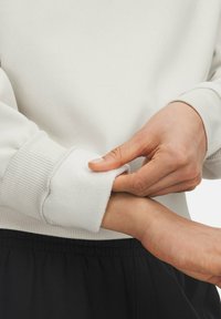 Felpa bianca con polsini a costine; il polsino interno è morbido e felpato. Una mano arrotola il polsino, rivelando la texture del tessuto. Pantaloni scuri visibili.