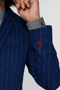 Blazer blu con motivo a righe, granchio ricamato sulla manica, dettaglio a quadretti grigi sul polsino e taschino obliquo sul petto.
