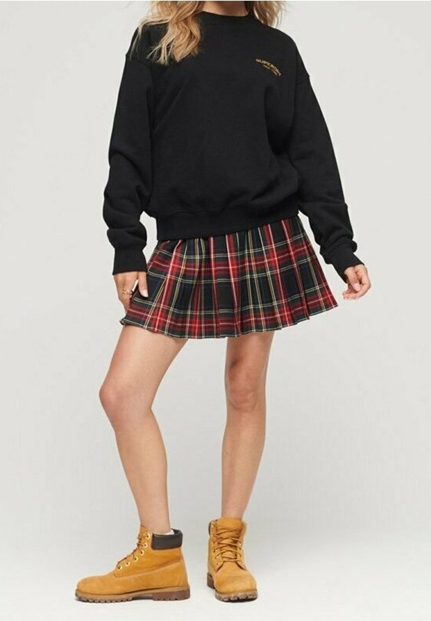 Personne portant un sweat-shirt noir, une jupe plissée à carreaux rouges et verts, et des bottes lacées beige, debout avec une jambe légèrement pliée.