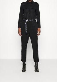 Combinaison noire à manches longues, avec deux poches poitrine et une taille cintrée. Association avec une ceinture noire comportant des graphiques contrastés. Bottines noires hauteur cheville.