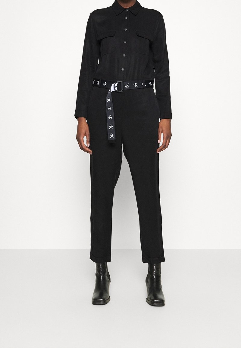 Combinaison noire à manches longues, avec deux poches poitrine et une taille cintrée. Association avec une ceinture noire comportant des graphiques contrastés. Bottines noires hauteur cheville.