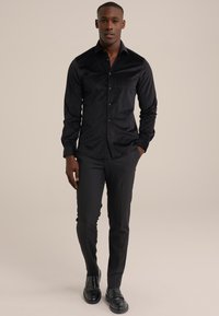 Camisa negra de botones con un sutil patrón texturizado, mangas largas y cuello, combinada con pantalones oscuros de corte ajustado y zapatos negros.