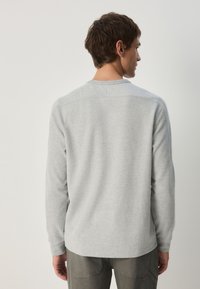 Hellgraues Langarmshirt mit gerippter Struktur, rundem Ausschnitt und gesteppten Details an den Schultern, getragen zu olivgrünen Hosen.