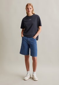 T-shirt bleu marine foncé à manches courtes avec du texte blanc, associé à un short en denim bleu et des baskets blanches avec des accents gris.