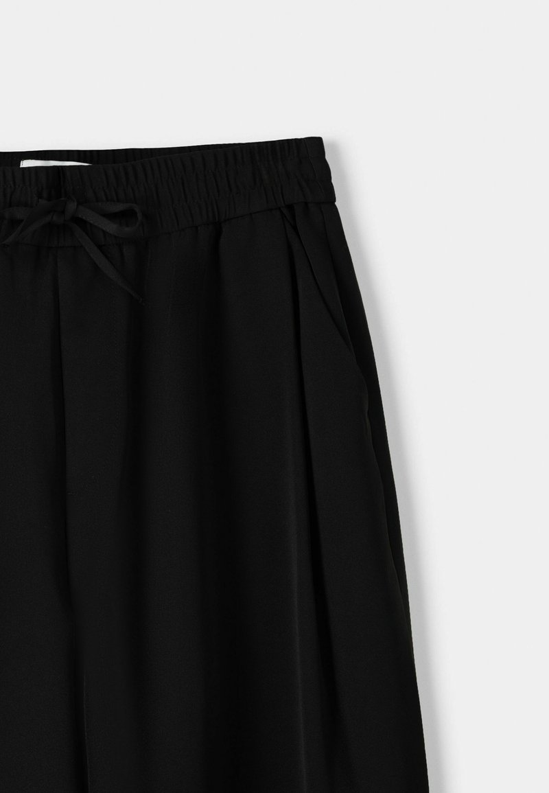 Czarne spodnie wykonane z gładkiego materiału, z elastyczną talią z możliwością regulacji, zakładkami z przodu i bocznymi kieszeniami.