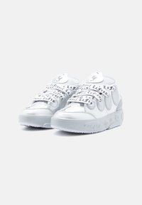 Adidași albi cu partea superioară din piele lucioasă, vârf texturat, limbă oversized și șireturi negre contrastante cu literele mărcii. Talpă groasă din cauciuc.