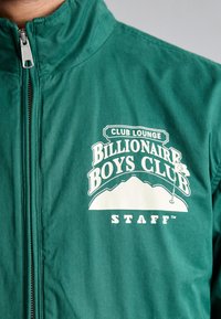 Grønn hettejakke med glidelås laget av glatt stoff, med hvit tekst og fjellgrafikk: "Billionaire Boys Club STAFF" på forsiden.