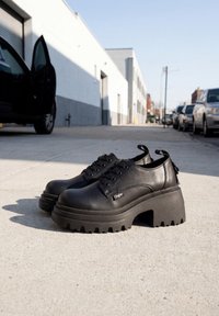 Chaussures à plateforme noires à lacets sur un trottoir ensoleillé en ville avec des voitures stationnées et un bâtiment en arrière-plan.