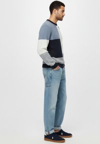 Man staat zijwaarts en draagt een kleurbloktrui in blauwtinten, lichtblauwe jeans met omslagen en marineblauwe sneakers met een wit logo.