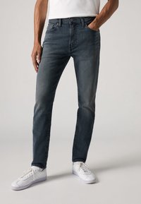 Muž v slim-fit tmavě modrých džínách a bílých teniskách, stojící s jednou rukou v kapse na jednobarevném pozadí.