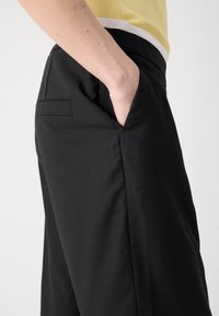 Pantalon noir avec une coupe ajustée, dotée de poches latérales et d'un tissu lisse et texturé. La taille est finie avec un design épuré et minimal.