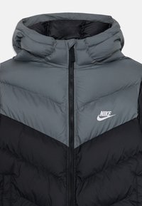 Veste matelassée en gris et noir, avec un col montant, une fermeture éclair et le logo Nike. Design matelassé avec un motif en chevrons et un matériau léger.