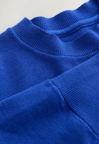 Jasnoniebieski sweter z teksturowanej tkaniny, z prążkowanym dekoltem i mankietami. Gładka część główna o luźnym kroju, podkreślająca prostotę w designie.