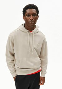 Beige hoodie met een zak aan de voorkant, trekkoorden in de capuchon en een geborduurd logo. Draagt een rood shirt eronder, gecombineerd met zwarte broek. Gladde textuur.